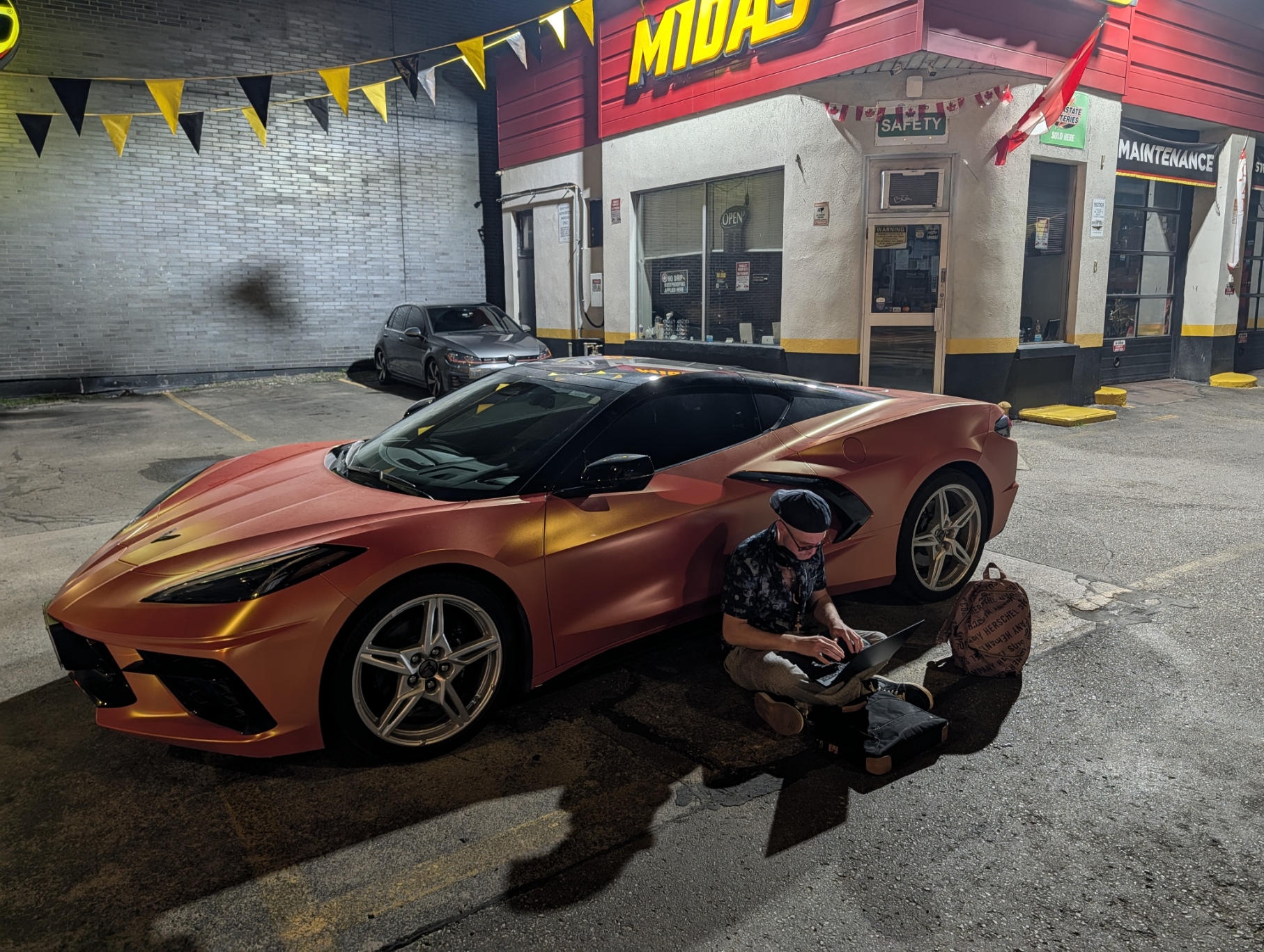 Man seated with a laptop beside an orange sports car outside the Midas shop at 401 Dixie Auto Mall at night